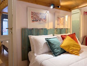 Ein Bett mit weißer Bettwäsche, farbenfrohen Kissen, einem hölzernen Kopfteil und zwei gerahmten Kunstwerken an der Wand.