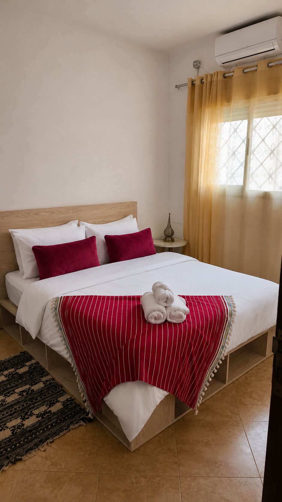 A neatly made bed with white and red bedding, a small bedside table with a lamp, and a window with curtains.