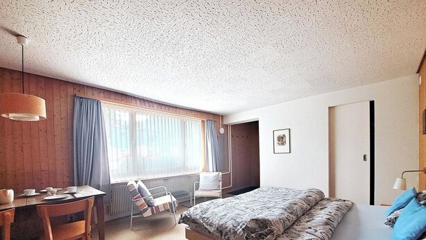 A hotel room with a bed, a desk, a chair, a radiator, and a window with curtains. - Chalet Willi-Parterre Appartement/Fewo, Dusche, WC, 2-Bettwohnung by Interhome (Arosa)