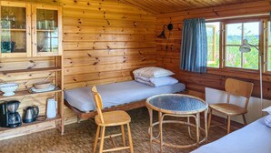 En stuga med en säng, två stolar, ett litet bord, ett skåp med glasdörrar och ett fönster med gardiner.