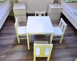 Una habitación pequeña y limpia con una mesa blanca y cuatro sillas, una mesita de noche y un tocador.