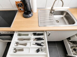 Una cocina con cafetera, dispensador de cápsulas de café, lavamanos y organizador de cajones con utensilios.