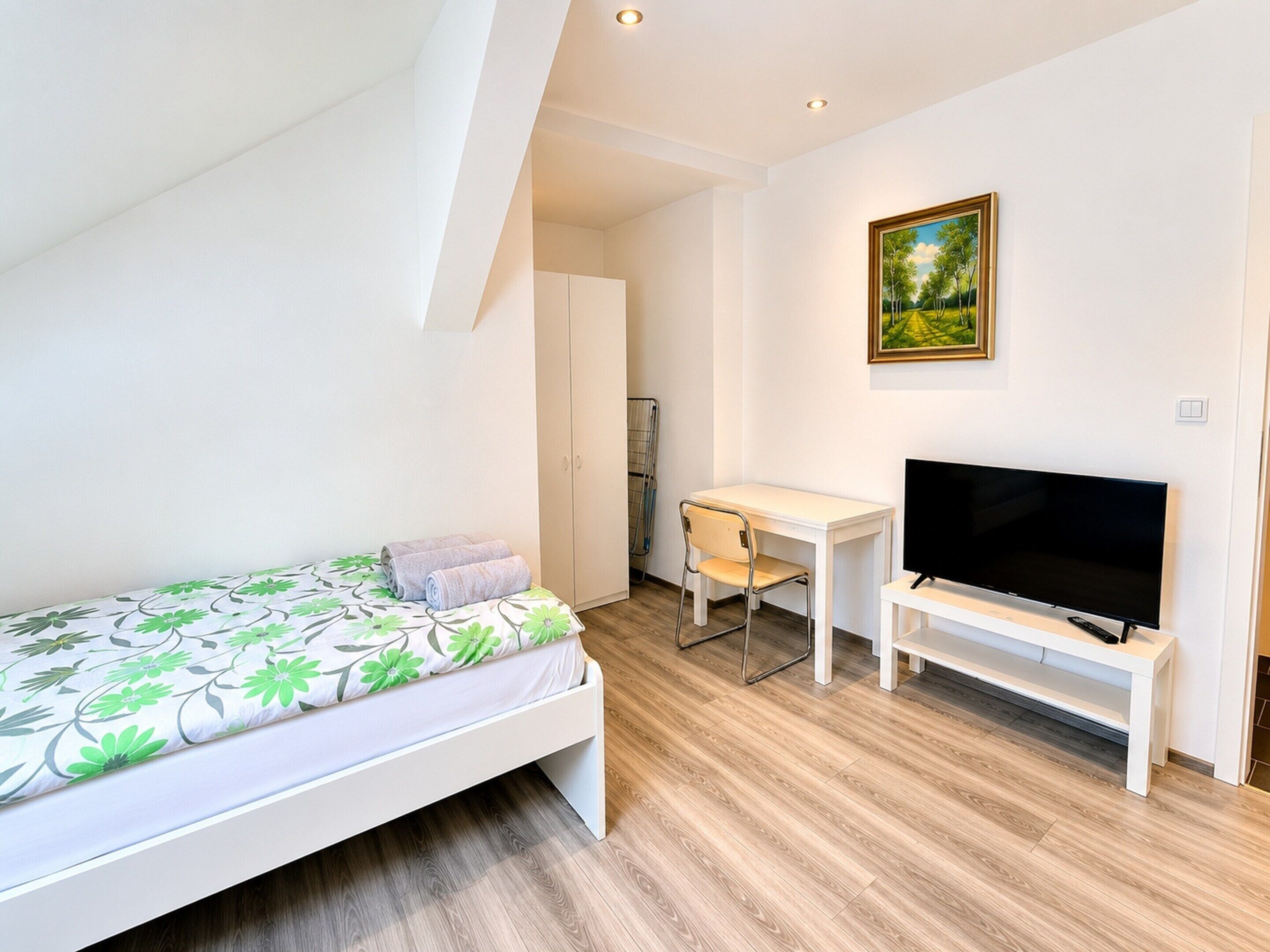 A bedroom with a bed, a desk, a chair, a TV, and a painting on the wall.