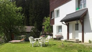 Een huis met een witte gevel, een houten terras en een kleine tuin met gazon en enkele stoelen.