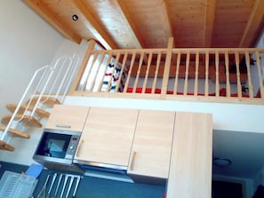 A loft bedroom with a wooden ceiling, a bunk bed, a kitchen area with a microwave, and a wooden staircase. - Haus Thöny Studio Appartement/Fewo,  , 2-Bettwohnung by Interhome (Arosa)