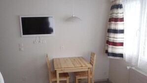 Een kleine kamer met een houten tafel en stoelen, een flatscreen televisie aan de muur bevestigd, en een raam met gestreepte gordijnen.