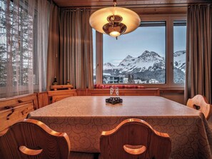 Ein Holztisch mit einem gemusterten Tischläufer, Holzstühle, eine Bank, eine Kristallleuchter und ein Blick auf schneebedeckte Berge durch große Fenster.