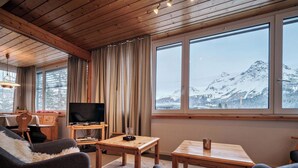 En hyggelig stue med tv, møbler af træ, et stort vindue med udsigt til et bjerglandskab og et spiseområde.