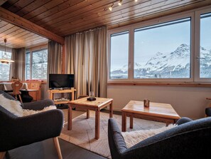 Ein gemütliches Wohnzimmer mit Fernseher, Holzmöbeln, einem großen Fenster mit Blick auf die Berge und einem Essbereich.