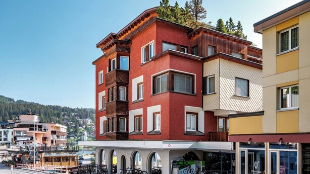 Un edificio de varios pisos con fachada roja, una tienda en la planta baja con bicicletas, y una vista de un paisaje montañoso al fondo.