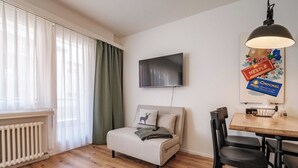 Una sala de estar moderna con un sofá, una mesa de comedor con sillas, un televisor de pantalla plana montado en la pared y un radiador debajo de la ventana.