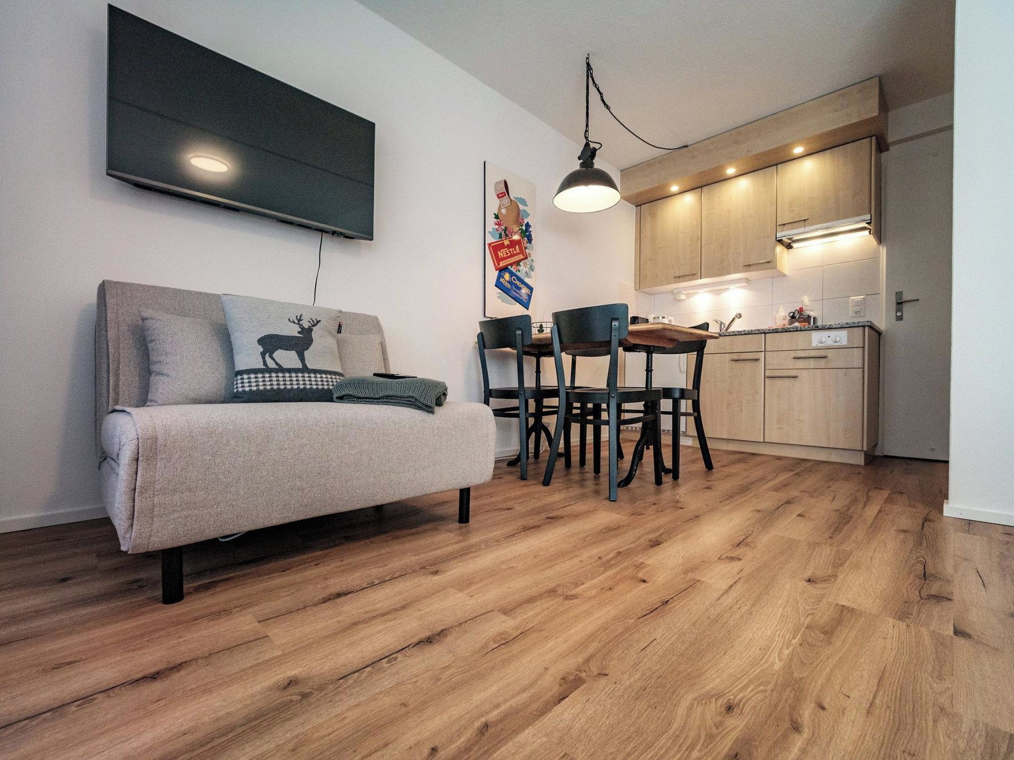 A modern apartment with a sofa, a dining table with chairs, a kitchen area, and a flat-screen TV mounted on the wall.