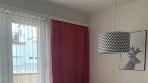 Een eetruimte met een witte tafel en vier stoelen, een kamerkleed met patroon, een rood gordijn en een wandlamp.