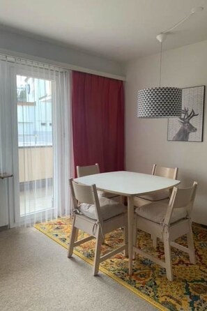 A dining area with a white table and four chairs, a patterned rug, a red curtain, and a wall-mounted lamp. - Paradies 04 Zweibettzimmer, Dusche oder Bad, WC, 2-Bettwohnung Paradies 04 by Interhome (Arosa)