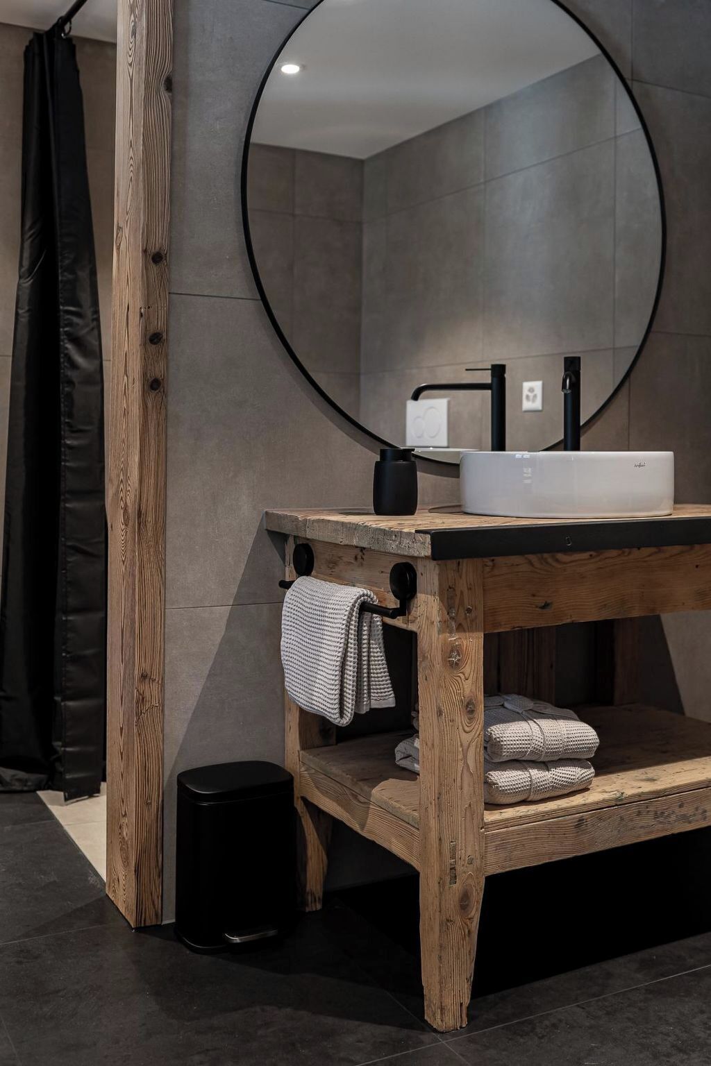 A bathroom with a wooden vanity, a round mirror, a black sink, and a towel rack.