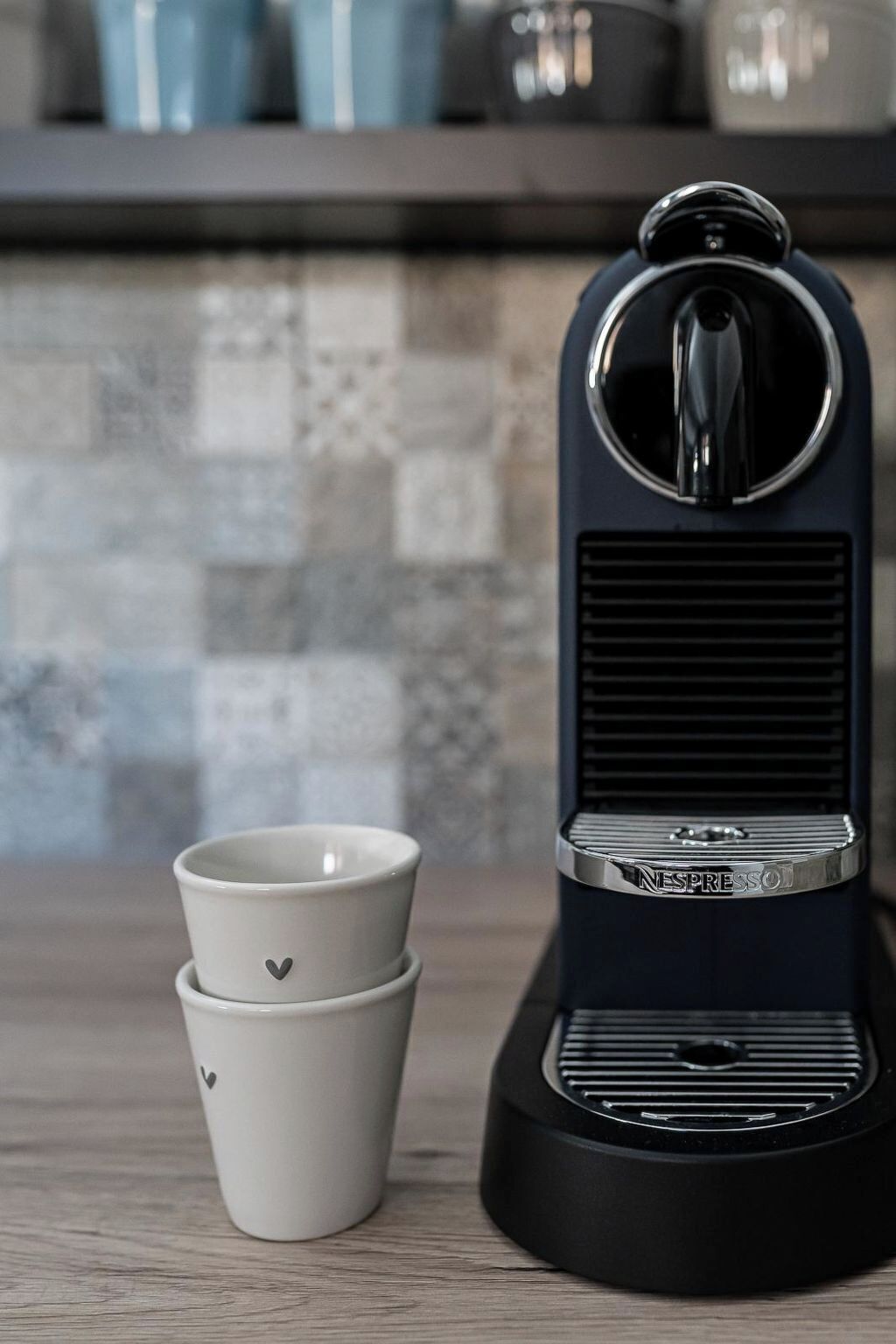 Una máquina de espresso negra con un logotipo de la marca en la parte frontal, dos tazas blancas apiladas con diseños de corazones, y un salpicadero de azulejos al fondo.