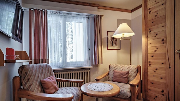 A cozy room with two wooden chairs, a small round table, a radiator, a window with curtains, a lamp, and a wooden wardrobe.