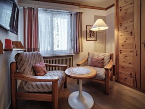 A cozy room with two wooden chairs, a small round table, a radiator, a window with curtains, a lamp, and a wooden wardrobe. - Paradies 206 Appartement/Fewo, Dusche, WC, 2-Bettwohnung 2-Bettwohnung by Interhome (Arosa)