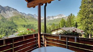 Varanda de madeira com mesa e cadeiras, com vista para uma paisagem montanhosa com uma casa e árvores.