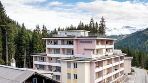 Ett hotell med flera våningar som heter ”PARADIES”, med balkonger, en parkeringsplats och en bergig bakgrund.