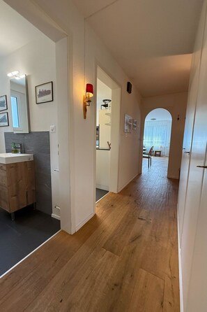 Un couloir doté d’un plancher en bois, une salle de bain avec un miroir et des tableaux encadrés, et une porte donnant sur une autre pièce.