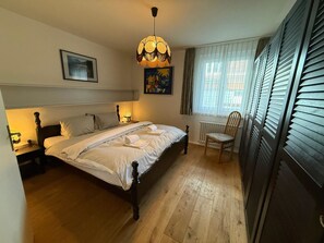 A bedroom with a bed, bedside lamps, a chair, a window with blinds, and a hanging light fixture. - Casa dUval by Arosa Holiday Appartement/Fewo, Dusche, WC, 6-Bettwohnung by Interhome (Arosa)