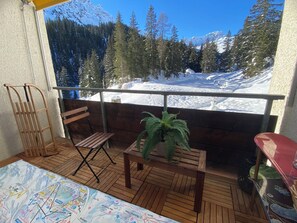 A balcony with a wooden floor, a wooden chair, a small table with a plant, and a view of snow-covered mountains and trees. - Rothornblick 6 Appartement/Fewo, Bad, WC, 4-Bettwohnung Rothornblick 6 by Interhome (Arosa)