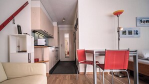 Un espacio habitable compacto con kitchenette, una zona de comedor con sillas rojas, un sofá y una mesita de noche con lámpara.