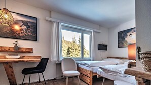 Ein Schlafzimmer mit Bett, Schreibtisch, Stuhl, Fenster mit Blick auf Bäume und einem Wand-Fernseher.