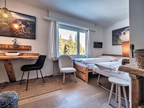 A bedroom with a bed, a desk, a chair, a window with a view of trees, and a mounted TV. - Paradies 603 Appartement/Fewo,  , 1-Bettwohnung 1-Bettwohnung by Interhome (Arosa)