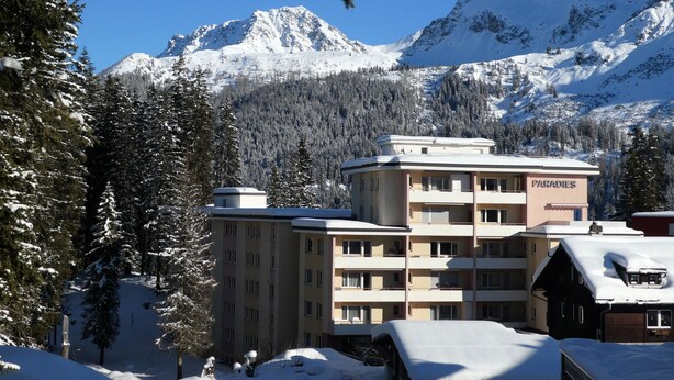 Ein mehrstöckiges Gebäude mit der Beschilderung 'PARADISES' im Hintergrund, umgeben von schneebedeckten Bäumen und Bergen.