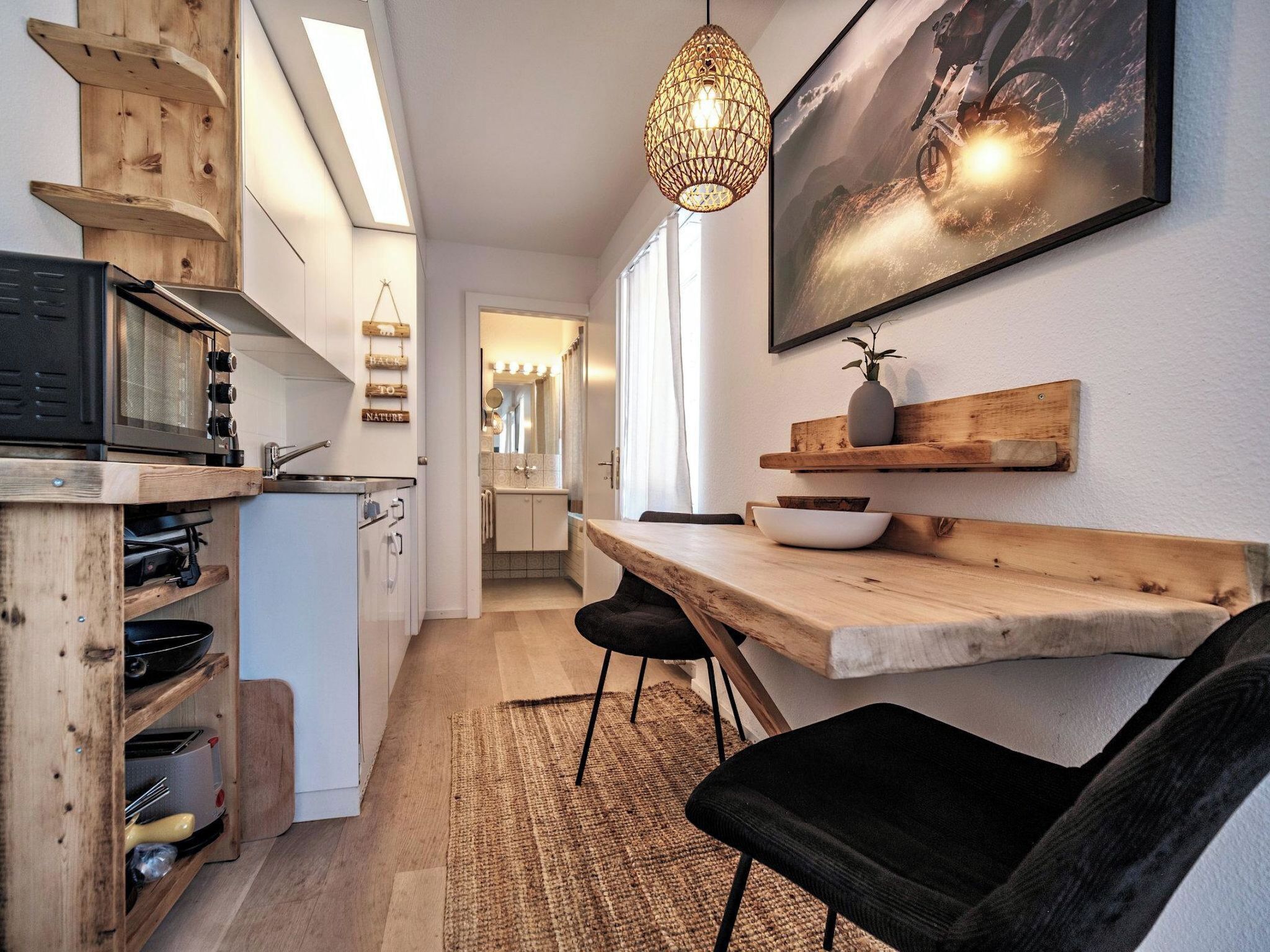 A compact kitchen with a microwave, wooden shelves, a hanging light fixture, and a dining area with a table and chairs.