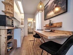 A compact kitchen with a microwave, wooden shelves, a hanging light fixture, and a dining area with a table and chairs. - Paradies 603 Appartement/Fewo,  , 1-Bettwohnung 1-Bettwohnung by Interhome (Arosa)