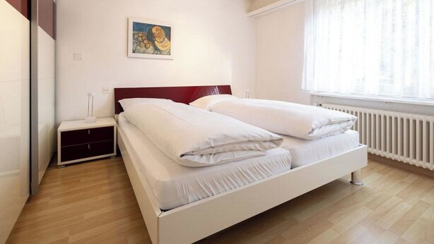 Un dormitorio con cama, mesita de noche, un cuadro y una ventana con cortinas.