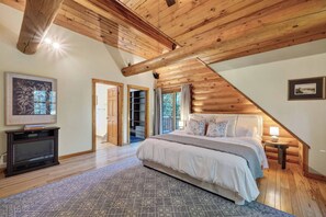 Un dormitorio con techo inclinado, vigas de madera, una cama con almohadas, un mueble para televisor y una alfombra.