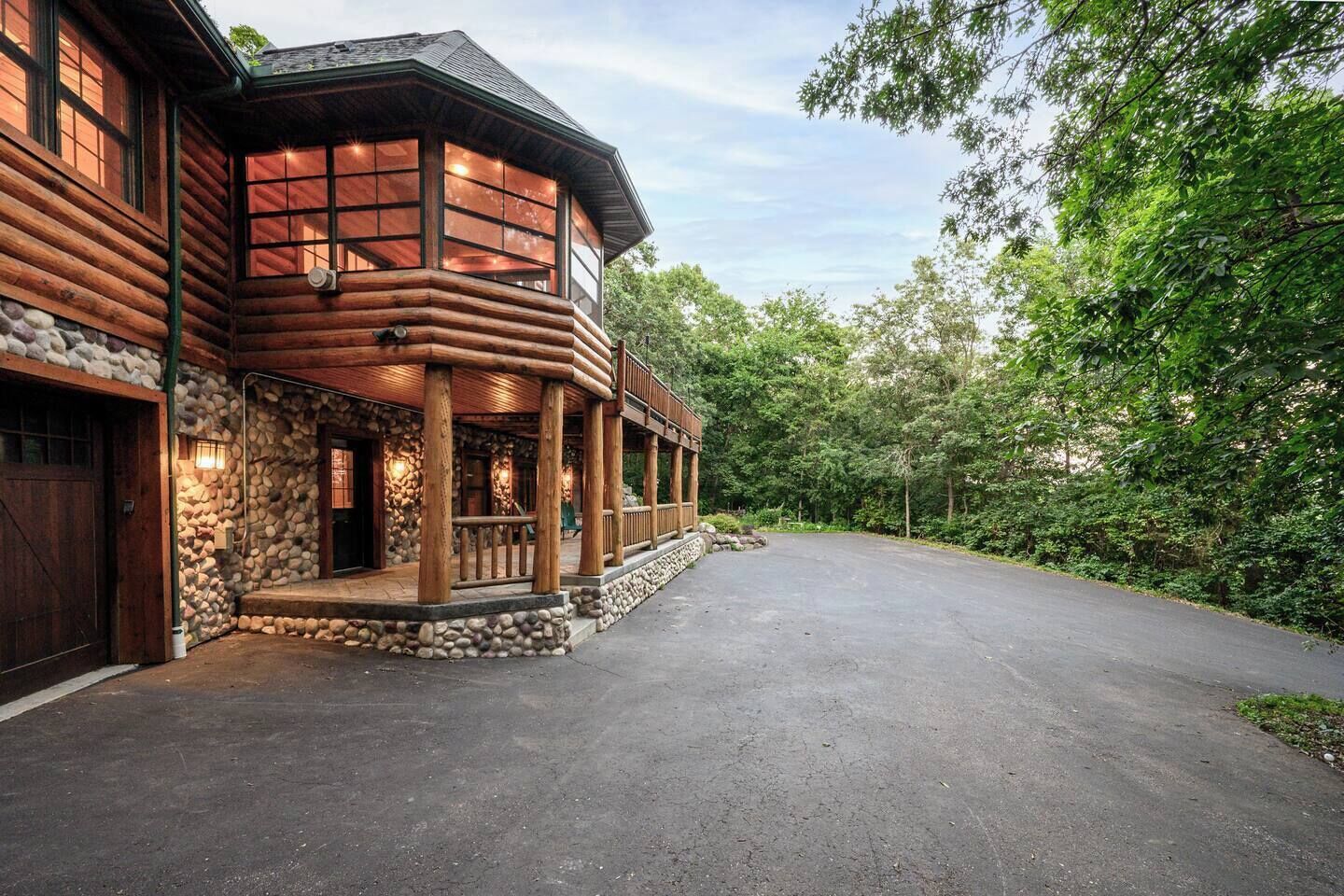 A log cabin with a stone foundation, a wooden porch, and a driveway leading to a garage.