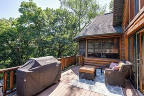 Una terraza de madera con una parrilla cubierta, mobiliario de mimbre y un ventanal que da a una habitación.