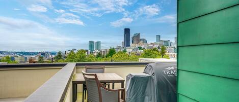A rooftop terrace with a table and chairs, a grill, and a city skyline view.