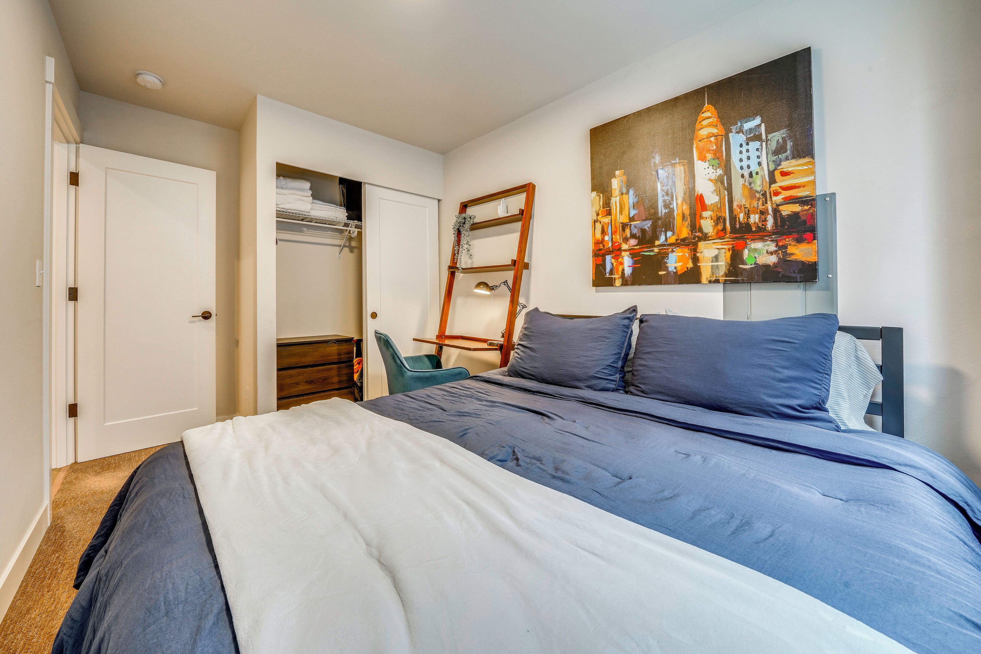 A bedroom with a bed, a closet, a chair, a ladder, and a cityscape painting on the wall.