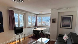 Ein modernes Wohnzimmer mit einer Couch, einem Glastisch, einem Fernseher auf einem Schrank, einem Essbereich mit Stühlen und großen Fenstern mit Blick auf Gebäude.
