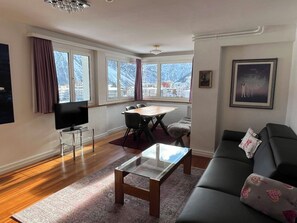 A modern living room with a sofa, a glass coffee table, a TV on a stand, a dining area with chairs, and large windows with a view of buildings. - Sunnybank / 3. St. Appartement/Fewo, Dusche und Bad, WC, 10-Bettwohnung by Interhome (Arosa)