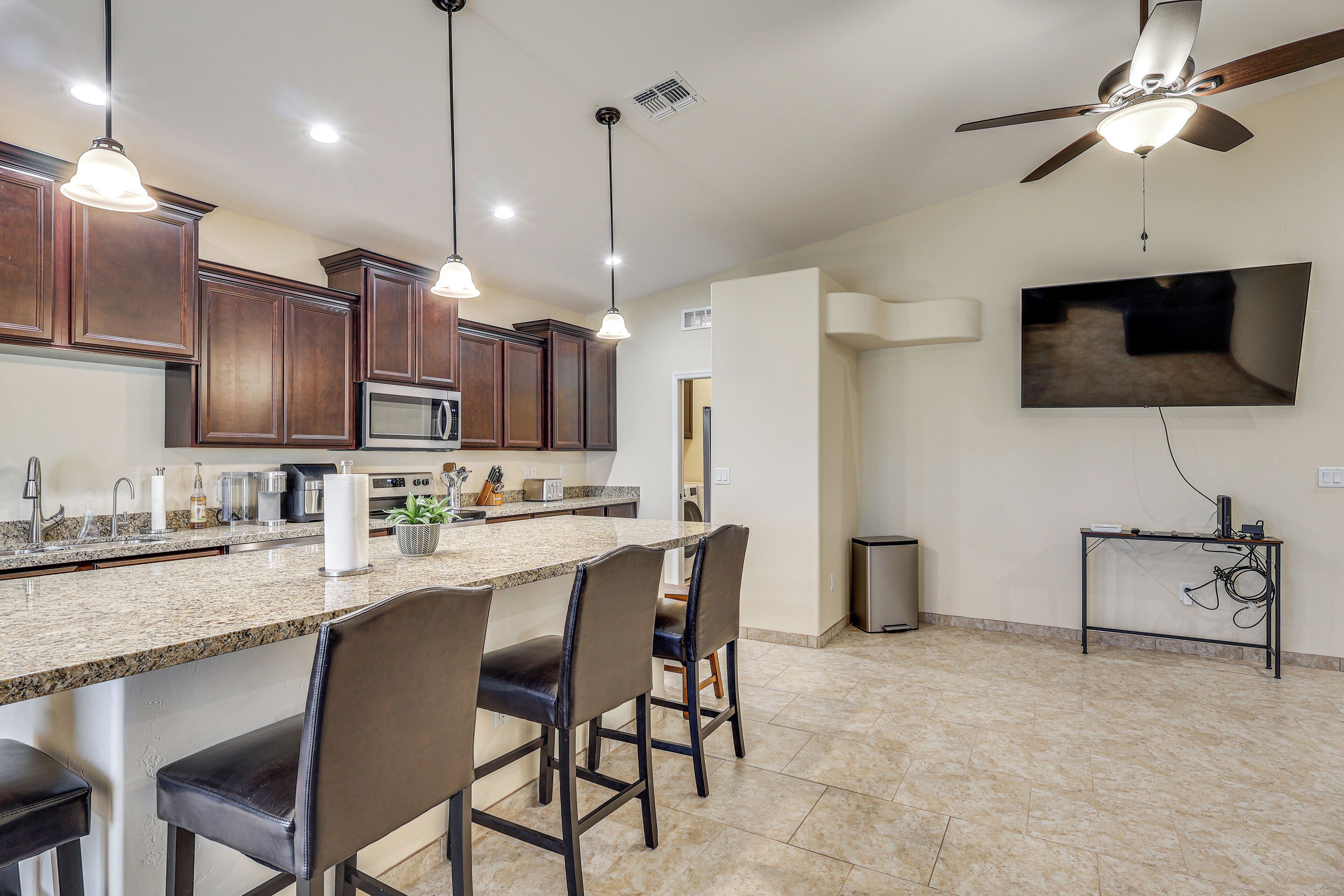 A kitchen with a large island, bar stools, wooden cabinets, a ceiling fan, and a mounted flat-screen TV.