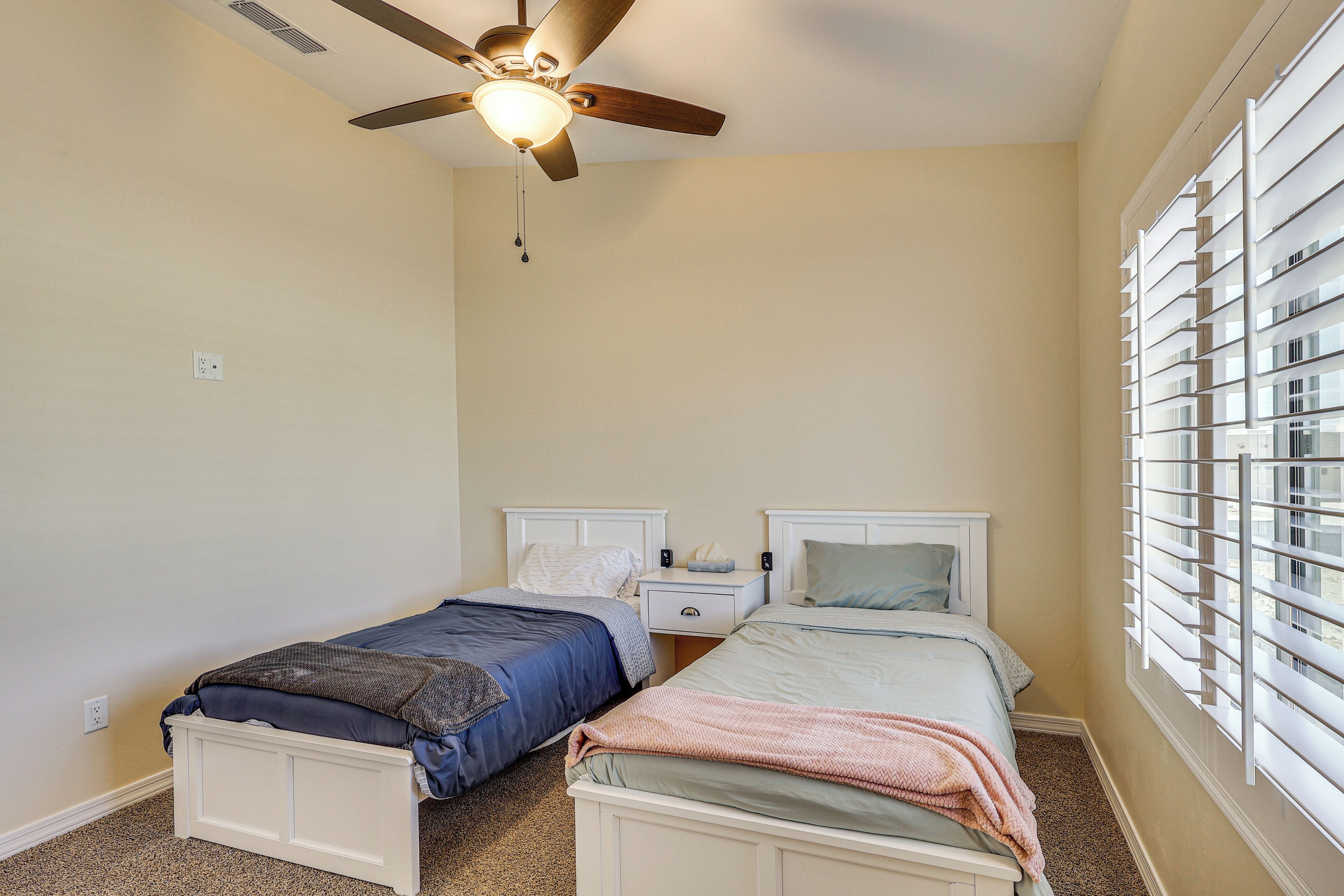 A bedroom with two single beds, a ceiling fan, a window with blinds, and a nightstand with a lamp.