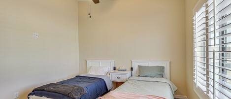 A bedroom with two single beds, a ceiling fan, a window with blinds, and a nightstand with a lamp.