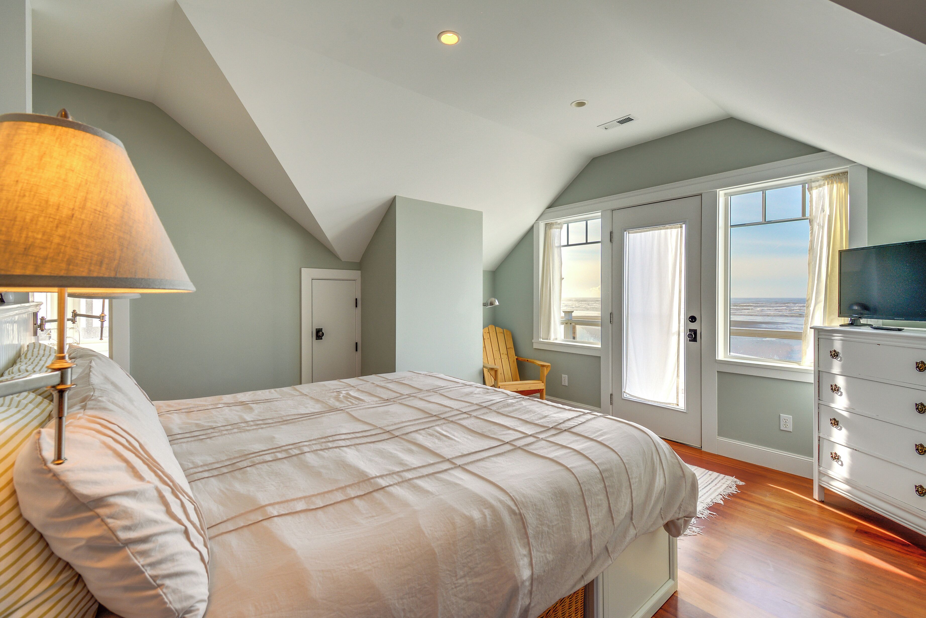 A bedroom with a large bed, a nightstand, a lamp, a dresser, and a view of the ocean through the windows.