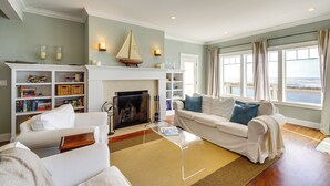 A living room with a fireplace, white furniture, a bookshelf, and a view of the ocean through large windows. - 4br/4ba Beachfront Retreat With Hot Tub and Sauna (Moclips)