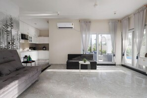 A modern living room with a grey sofa, a small round table with a plant, and a kitchen area with white cabinets and a built-in stove.