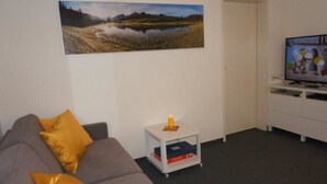A living room with a grey sofa, a small white side table with a candle, and a flat-screen TV on a white stand. - Paradies 505 Appartement/Fewo,  , 2-Bettwohnung 2-Bettwohnung by Interhome (Arosa)