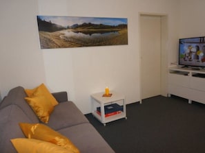 Ein Wohnzimmer mit einem grauen Sofa, einem kleinen weißen Beistelltisch mit Kerze und einem Flachbildfernseher auf einem weißen Lowboard.