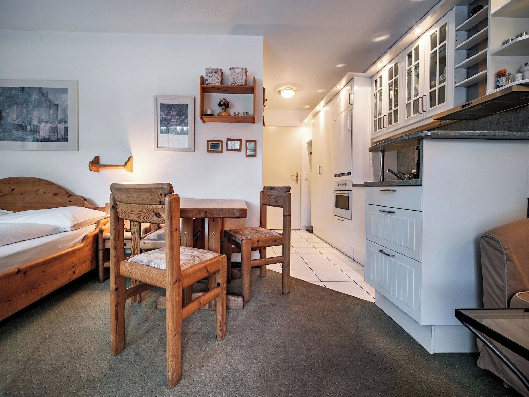 Een knus ingerichte kamer met een bed, een houten eettafel met stoelen, een kitchenette en een klein plankje met decoratieve spullen.